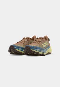 Sapatos de corrida em trilhos com parte superior em malha em tons terrosos, uma entressola com padrão azul e verde, e uma sola externa de borracha robusta para tração.