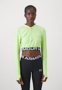 Fényes zöld, hosszú ujjú crop top, cipzáras gallérral és logóval. Fekete, magas derekú leggingsszel párosítva, márkás derékkal.