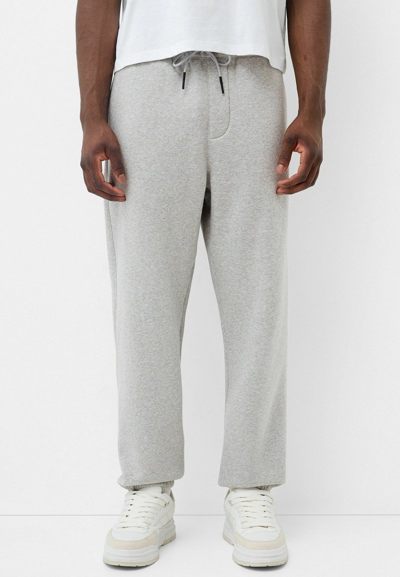 Graue Sweatpants mit elastischem Bund und Kordelzug, ausgestattet mit Seitentaschen. Tragen Sie sie mit weißen Turnschuhen, das Material wirkt weich und lässig.