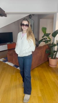 Valge tõmblukuga fleece-jakk roosa topi peal, tumelõhnavad sinised alt laienevad teksad, hallid saapad, suured tumedad päikeseprillid, seisab moodsas siseruumis.