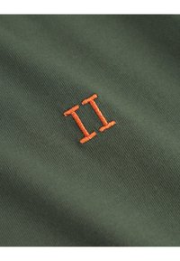 Zeleni tekstil z teksturirano površino, s poudarjenim oranžnim vezenim številom "II" v minimalističnem dizajnu.