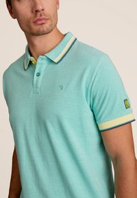 Lichtblauwe polo gemaakt van gestructureerde stof, met een contrasterende kraag en manchetten met gele en marineblauwe strepen, en een klein logo aan de voorkant.