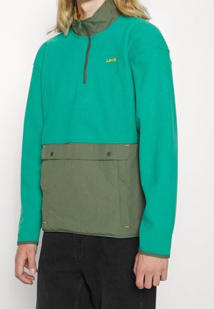 Person trägt einen Fleece-Pullover mit Viertelreißverschluss in Blaugrün und Olivgrün, mit Frontklappentasche und Levi's-Logo auf der Brust.