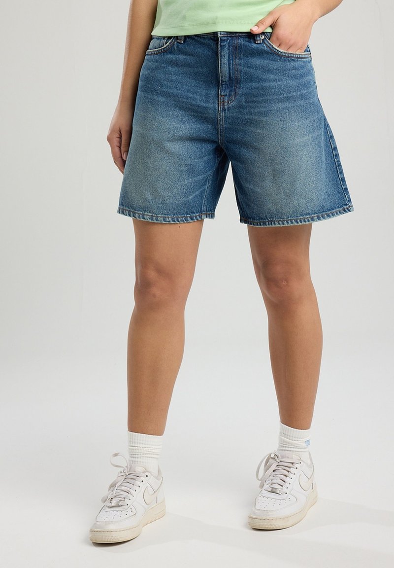 Pantalones cortos de mezclilla en azul claro, con un ajuste relajado, un ligero desgaste y un corte recto. Combinados con zapatillas blancas y calcetines texturizados.