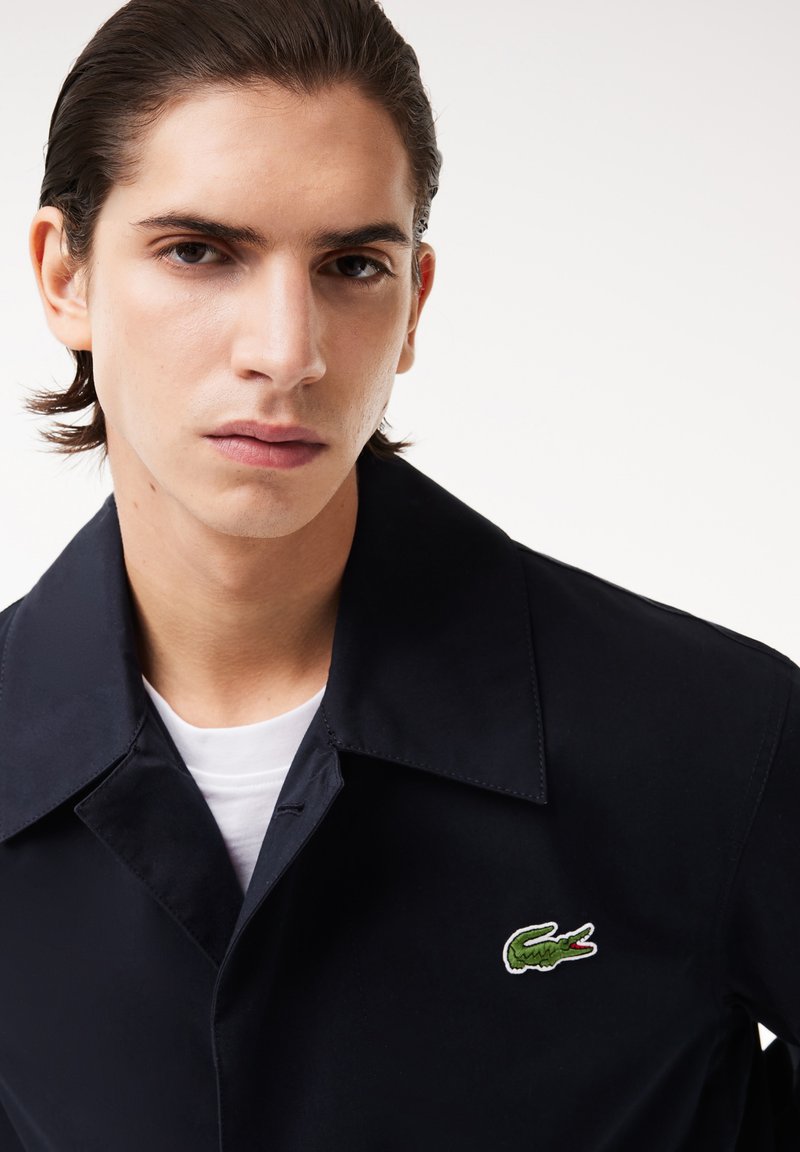 Abrigo navy con un cuello estructurado, presentando un logotipo de cocodrilo verde bordado en el lado izquierdo del pecho. Camiseta blanca lisa visible.