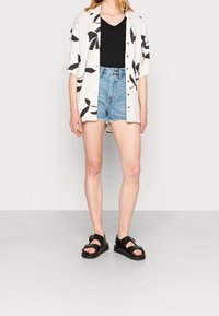 Camisa de manga corta de color claro con patrón de hojas negras, sobre una blusa negra de cuello en V. Pantalones cortos de mezclilla azul con dobladillos deshilachados y sandalias negras.