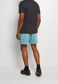 Man i svart mönstrad t-shirt, ljusblå shorts, svarta strumpor och svarta Nike-sneakers, står mot en enkel vit bakgrund.