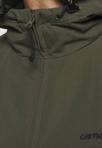 Olivgrüne Jacke mit hohem Kragen, verstellbaren Kordelzügen und sichtbaren Nähten. Mit Logo-Stickerei in dunkler Farbe an der unteren Seite.