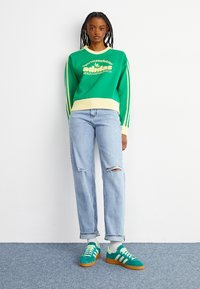 Zelená mikina se žlutými akcenty a bílým textem; modré roztrhané džíny; tyrkysové semišové tenisky s bílými pruhy; stylizováno s bílými ponožkami.