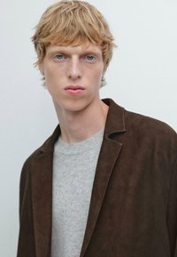 Bruine suède blazer over een lichtgrijs gebreid sweater; close-up van een model met licht haar, blauwe ogen en een neutrale achtergrond.