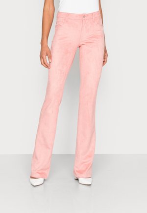 Mujer con pantalones rectos de ante rosa claro y zapatos blancos de punta afilada, de pie frente a un fondo liso gris claro.