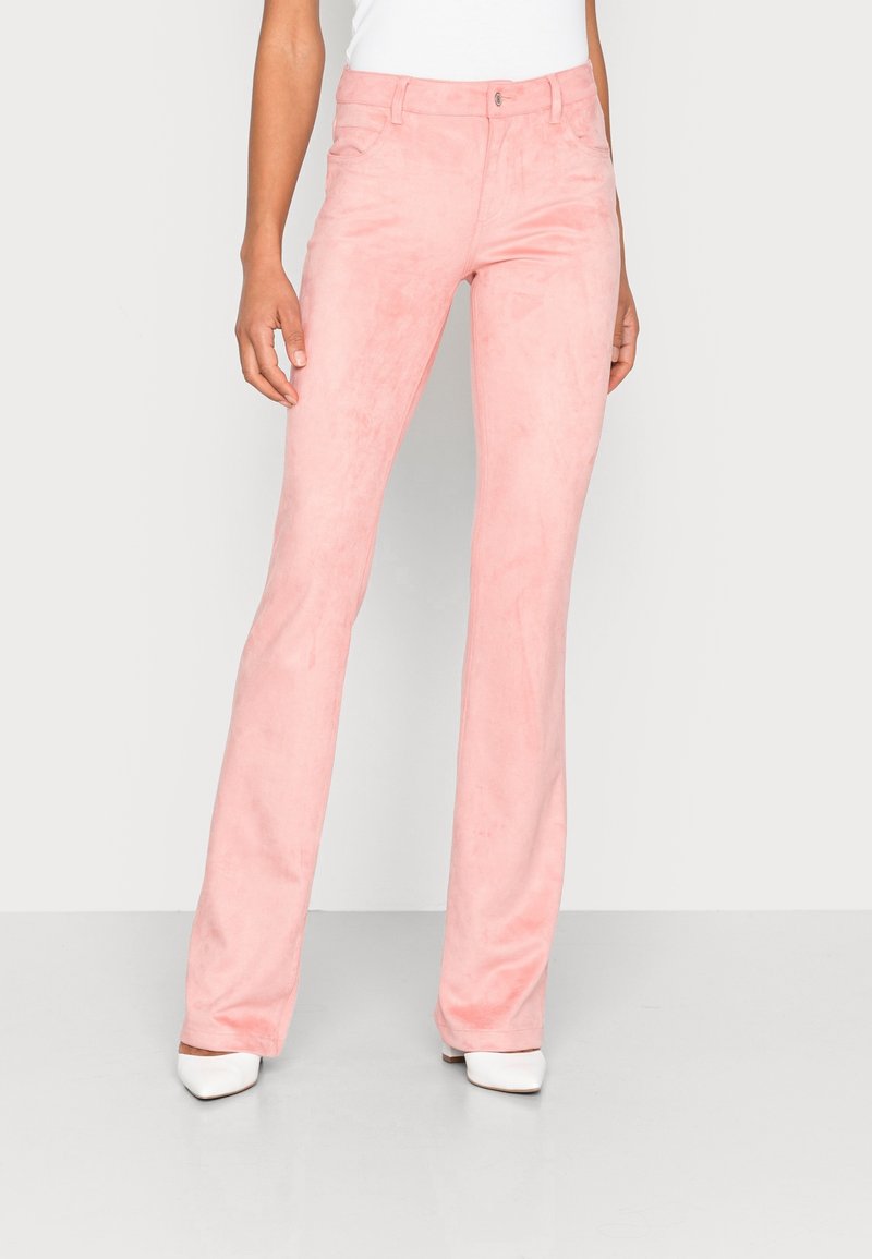 Femme portant un pantalon en daim rose clair à jambes droites et des chaussures blanches à bout pointu, debout devant un fond uni gris clair.
