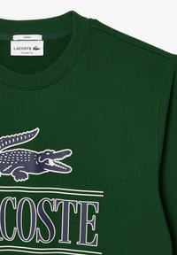 Sweatshirt en coton vert avec un col rond, arborant une marque Lacoste blanche et un logo crocodile navy sur le devant. Design à coupe classique.