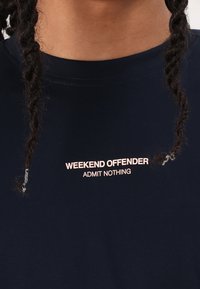 Marineblå t-skjorte med korte ermer, med teksten "WEEKEND OFFENDER" og "ADMIT NOTHING" i lys rosa på brystet. Laget av bomull.