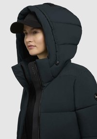 Geacă puffer închisă, cu o glugă mare, realizată dintr-un material mat, având un fermoar negru și un patch cu logo pe mânecă.