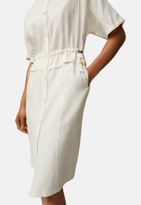 Abito camicia button-up di colore crema chiaro, con una vestibilità rilassata, vita arricciata e tasche laterali. Presenta un tessuto morbido e maniche corte.