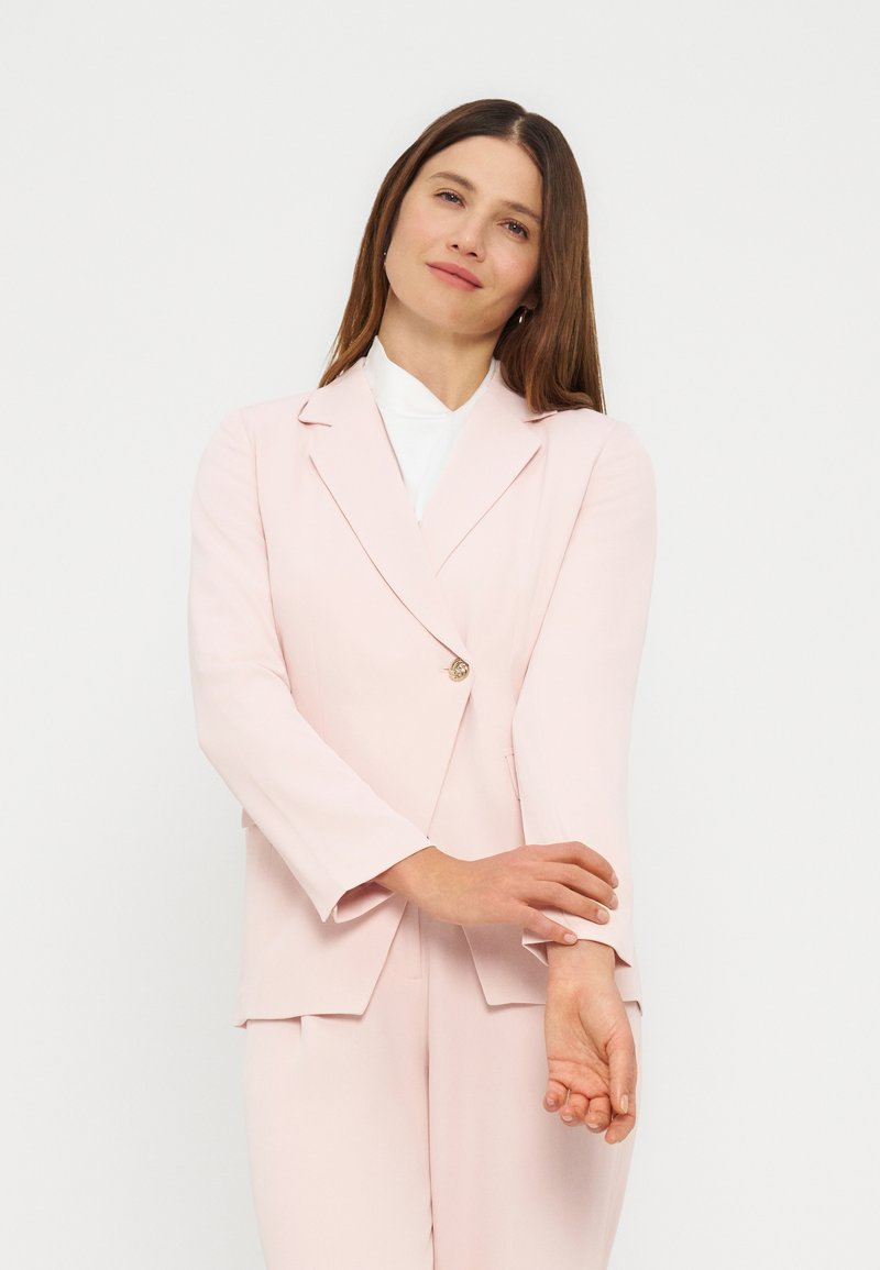 Γυναίκα με μακριά καστανά μαλλιά, φορούσα ένα ανοιχτό ροζ κοστούμι και άσπρο τοπ, ρυθμίζει το αριστερό της μανίκι, στέκεται μπροστά σε απλό λευκό φόντο.