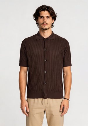 Man met donker haar draagt een bruin gebreid shirt met korte mouwen en knopen en beige broek, staand tegen een effen lichte achtergrond.