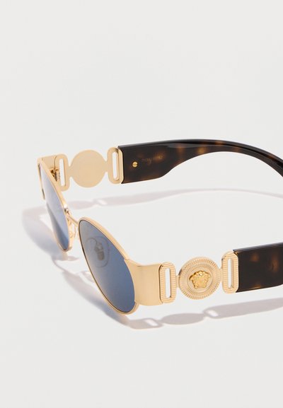 Γυαλιά ηλίου με χρυσό σκελετό και σκούρους φακούς, με φαρδιές βραχίονες σε σχέδιο χελώνας και κυκλικό έμβλημα με διακοσμητικό κεφάλι λέοντα.