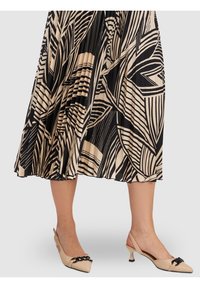 Jupe plissée noire et crème avec des motifs géométriques, assortie à des escarpins beige à talon, ornés d'une chaîne noire décorative.