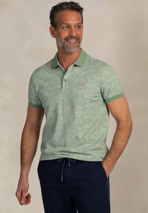 Man die lacht, gedragen een lichtgroen polo met patroon en kraag, en marineblauwe broek met trekkoord, staand tegen een effen achtergrond.