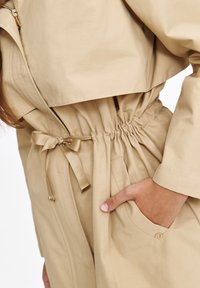 Jasno beżowa kurtka z talią, zamkiem błyskawicznym i kieszeniami naszytymi. Wykonana z gładkiego materiału o lekko połyskującym wykończeniu.