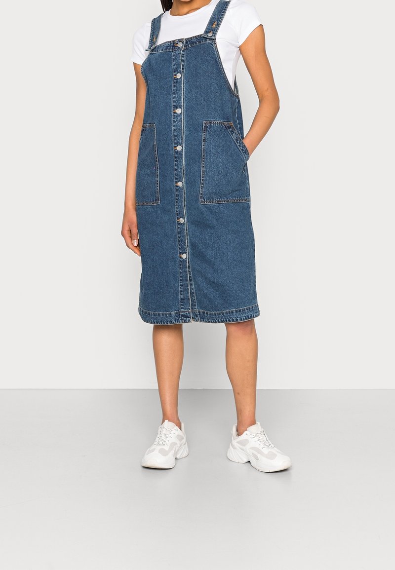Femme portant une robe salopette en denim bleu à boutons sur un t-shirt blanc avec des baskets blanches, debout avec une main dans la poche.