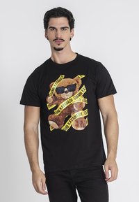 Camiseta de algodón negra con gráfico de un oso usando gafas de sol, rodeado de cinta de marca amarilla con texto repetido, mangas cortas y cuello redondo.