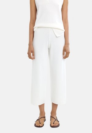 Białe, szerokie spodnie dresowe o gładkiej teksturze i długości do połowy łydki, zestawione z białym topem bez rękawów i brązowymi płaskimi sandałami.