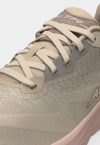 Κοντινό πλάνο μπεζ αθλητικού παπουτσιού με υφασμάτινο διχτυωτό υλικό, σταυρωτά κορδόνια και διακριτικό ροζ λογότυπο στη γλώσσα και στο πλευρό.