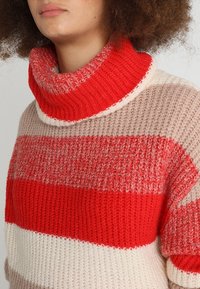 Pull en maille avec un col roulé haut rouge, présentant des rayures horizontales en rouge, beige et crème. Tissu texturé avec une coupe ample.