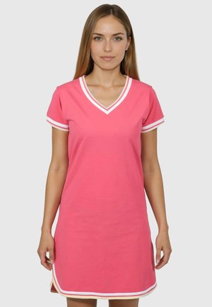 Donna che indossa un vestito rosa a maniche corte con bordi a righe bianche sullo scollo a V, le maniche e l'orlo, in piedi contro uno sfondo semplice.