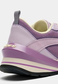 Zapatilla deportiva morada con parte superior de malla texturizada, detalles en cuero suave y suela gruesa. Características destacadas incluyen una lengüeta del talón morada y un logotipo.