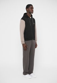 Sudadera negra con cremallera y mangas beige, con un logotipo frontal. Combinada con pantalones grises y zapatillas blancas, mostrada en un modelo de pie.