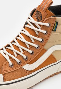 Høye sneakers med brunt semsket skinn og kanvasmateriale, hvite snørebånd, brune øyeletter og en tan-design. Har en hvit sidestrek og logo.