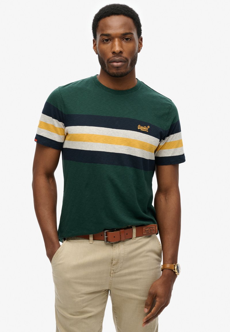 Grøn t-shirt med korte ærmer og navy, grå og sennepsgule striber; har et broderet logo på brystet og bæres med lysebeige bukser.