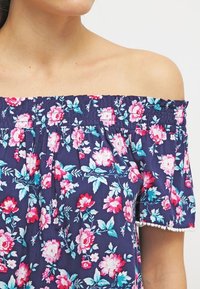 Top floreale scollato in blu navy con fiori rosa e turchesi, caratterizzato da bordi arricciati e un arricchimento decorativo sulle maniche.