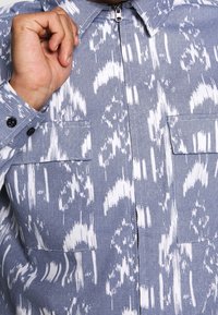 Blue and white patterned shirt with a zip closure, two chest pockets, and button accents on the cuffs. Textured fabric with a modern design.