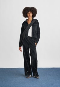 Μαύρο σατέν φόρεμα άθλησης με τζάκετ που κλείνει με φερμουάρ και ελαστικές μανσέτες, παντελόνι με ελαστική μέση, συνδυασμένο με τσάντα χιαστί.