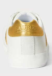 Λευκό αθλητικό παπούτσι με χρυσή δερμάτινη λεπτομέρεια στη φτέρνα, με υφή και ανάγλυφο λογότυπο. Ομαλή συνολική σχεδίαση.