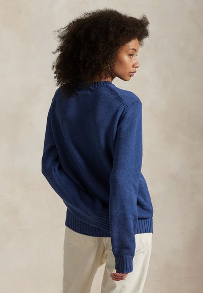 Frau mit lockigem Haar, seitlich gewandt, trägt einen blauen Strickpullover und helle Hosen vor einem neutralen Hintergrund.