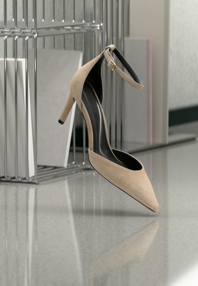 Μπεζ γοβάκι με τακούνι και λουράκι στον αστράγαλο, αιωρούμενο μπροστά από μεταλλική βάση για περιοδικά πάνω σε αντανακλαστικό πάτωμα.