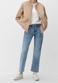 Persona vestida con una chaqueta bomber beige, camiseta blanca, jeans de cintura alta azul claros con ligera degradación y zapatillas blancas, de pie contra un fondo blanco.