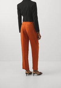 Black cropped blazer paired with orange trousers. Trousers feature a smooth texture, straight cut, and back pockets. Black pointed heels complete the outfit.
