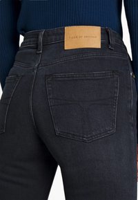 Black denim jeans with a smooth texture, five-pocket design, and a tan leather patch labeled "TIGER OF SWEDEN" on the waistband.