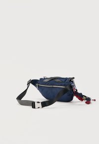 Riñonera azul marino con cremallera plateada, correa negra ajustable y un llavero de cuerda multicolor adjunto sobre un fondo claro.