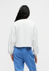 Κοντό, ανοιχτό γκρι φούτερ με μακριά μανίκια και στρογγυλή λαιμόκοψη, σε συνδυασμό με μπλε φόρμες, εμφανίζεται από την πίσω πλευρά.