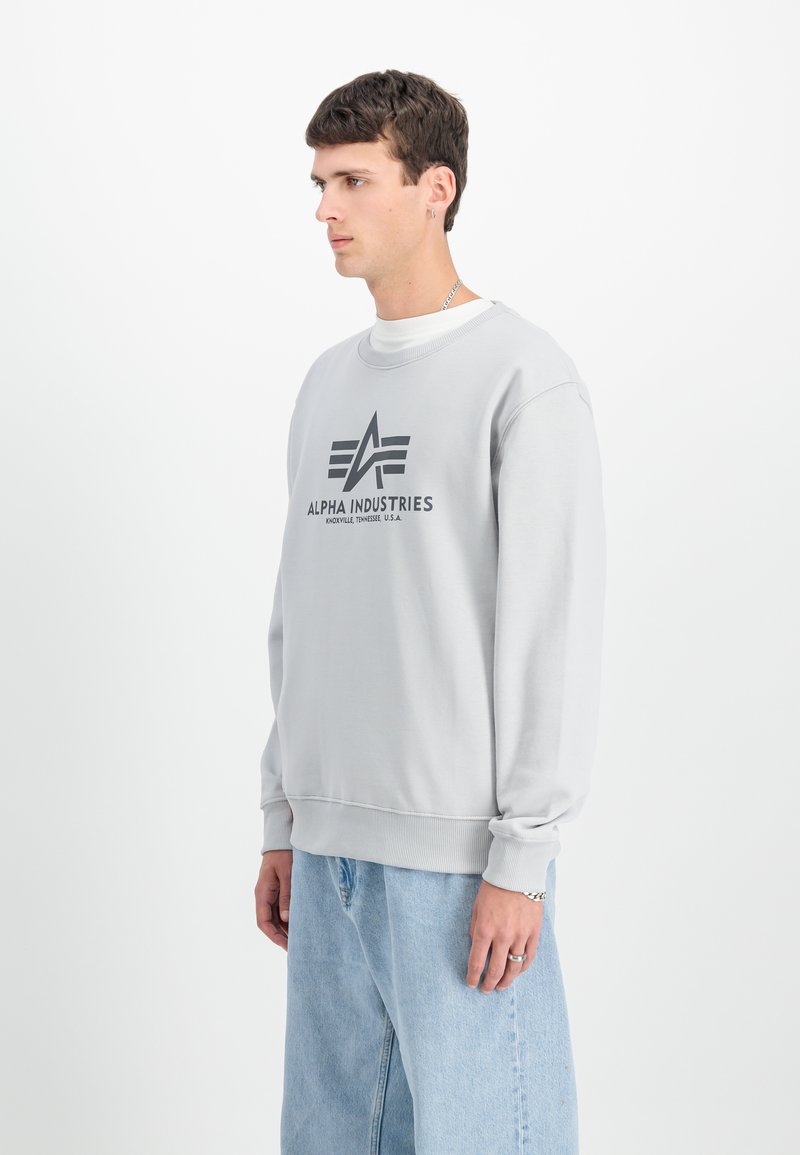 Mladý muž otočený doľava, oblečený v svetlošedej mikine Alpha Industries a svetlomodrých džínsoch na jednoliatom bielom pozadí.