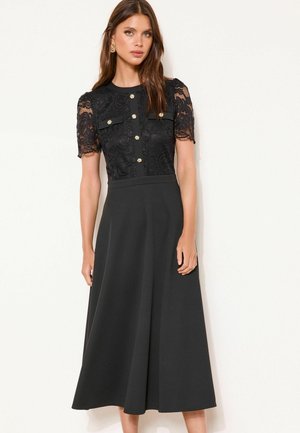 Nő fekete ruhát visel csipkés rövid ujjakkal, arany gombokkal az elején és A-vonalú szoknyával, fehér háttér előtt áll.