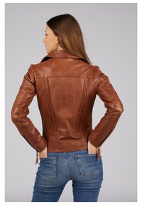 Braune Lederjacke mit kunstvollen Nähten entlang der Rückennähte, strukturiertem Kragen und Reißverschlüssen an den Ärmelabschlüssen. Kombiniert mit blauen Jeans.